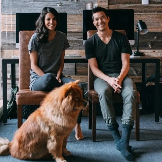 Candid photo of two young professionals sitting with a golden retriever in a modern workspace, wood-paneled wall and desktop monitors in the background.